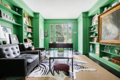 A home office with green built-in bookshelves, two dark leather armchairs, a glass-top coffee table with a small ottoman, and a desk facing a window.