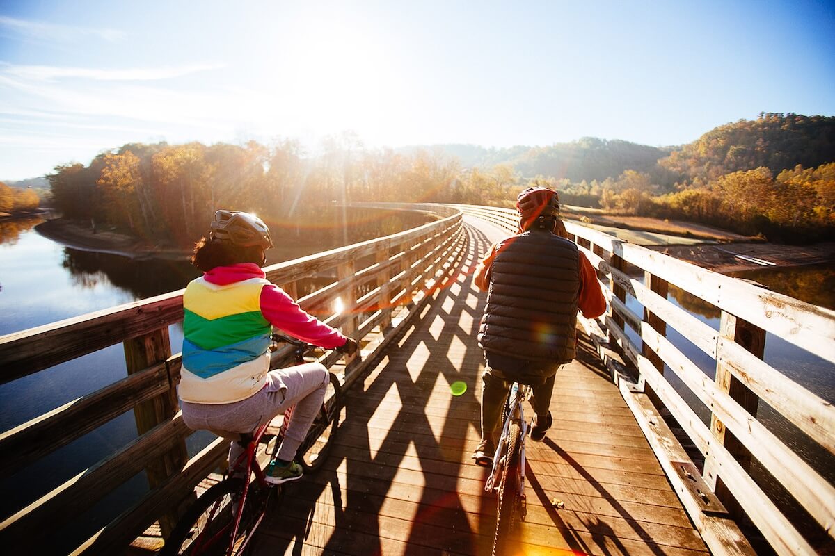 Two people wearing helmets ride bicycles on a wooden bridge in small town Virginia under a clear sky. The sun is shining, and trees with autumn foliage are visible in the background.