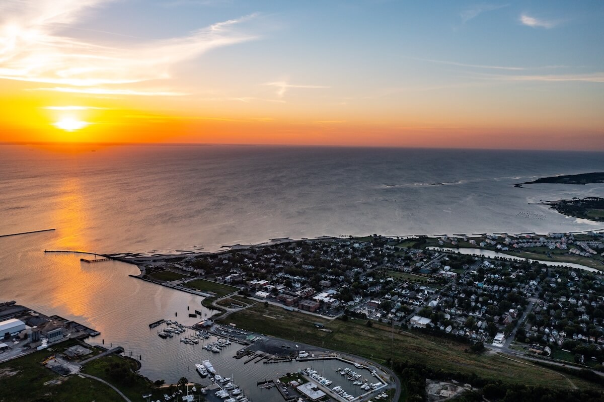 Aerial view of a small Virginia coastal town at sunset, with a vibrant orange sky, calm sea, and visible pier. Residential areas and the marina grace the foreground.