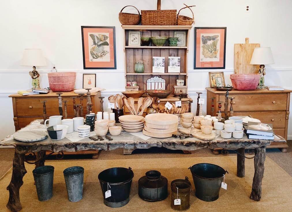 A rustic store display in small-town Virginia features a wooden table with various bowls, plates, and kitchenware. In the background, two wooden cabinets adorned with decorative items and baskets capture the quaint charm of this charming community.