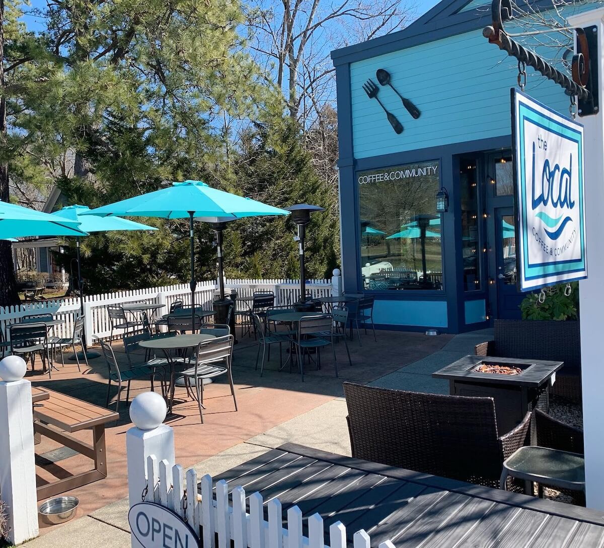 Outdoor cafe with turquoise umbrellas and black chairs, surrounded by a white picket fence in small town Virginia. A blue building displays utensils decor and signage reads "coffee & community.