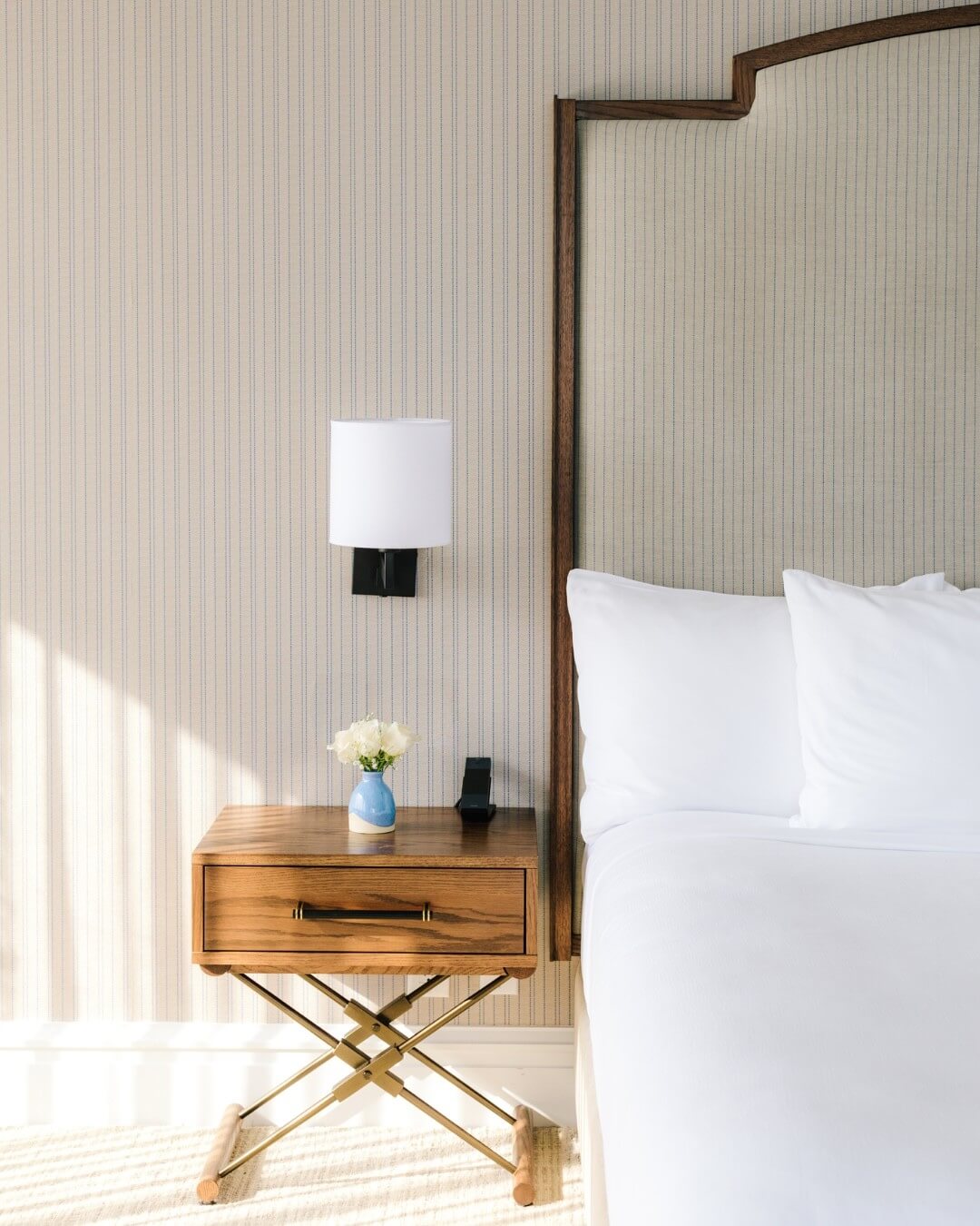 A neatly made bed with white pillows is next to a wooden nightstand, which evokes the cozy charm of small-town Virginia, featuring a small lamp, a vase of white flowers, and a phone on top.