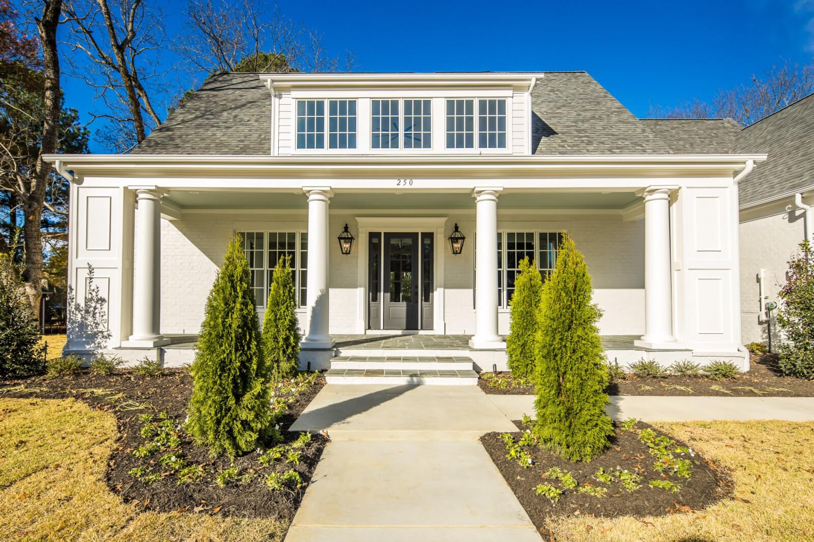 A white two-story Magnolia Homes design features columns, large windows, and two lanterns flanking the entrance. A path, bordered with trimmed shrubs and lush grass, guides you to the front door.