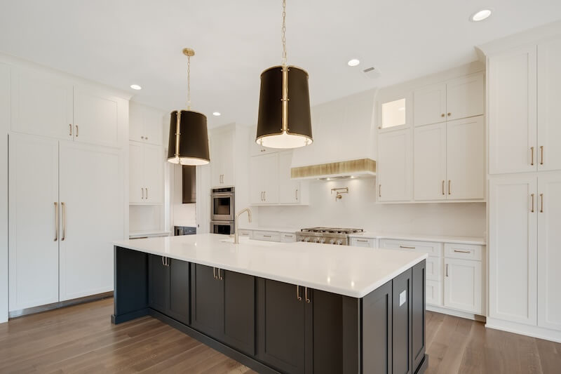A modern kitchen with a large island, white cabinets, stainless steel appliances, two black pendant lights, and wooden flooring.