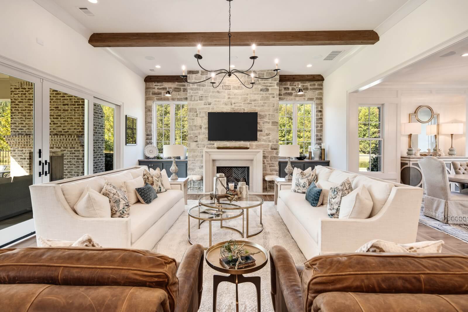 A spacious living room designed by Magnolia Homes features two white sofas, a central coffee table, and a fireplace. Large windows provide natural light, while a chandelier hangs from the ceiling with exposed wooden beams.