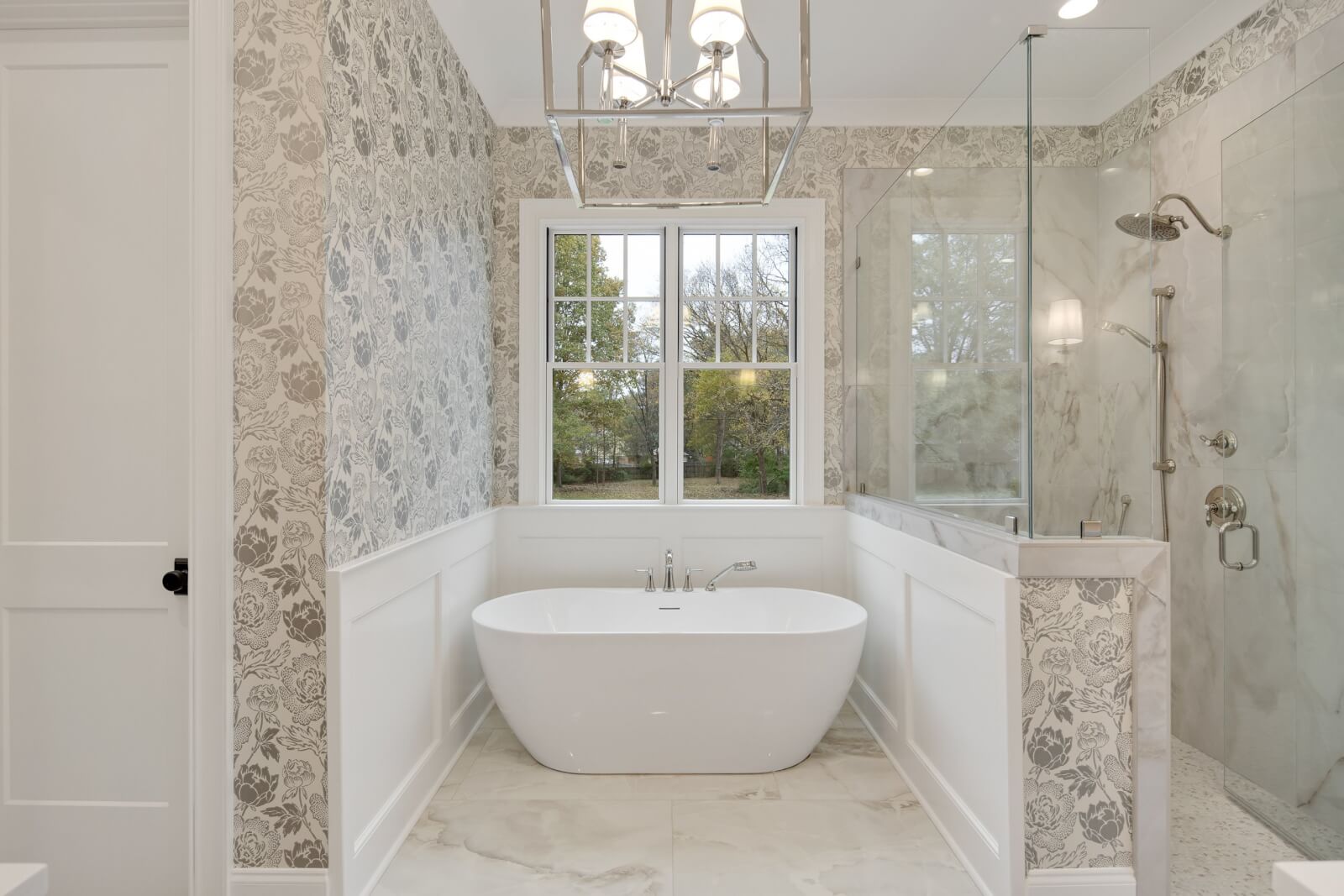 Elegant bathroom with a freestanding bathtub, glass shower, and large window. The floral wallpaper adds charm to the home design, while a modern chandelier overhead enhances the sophisticated interior decorating inspired by Magnolia Homes.