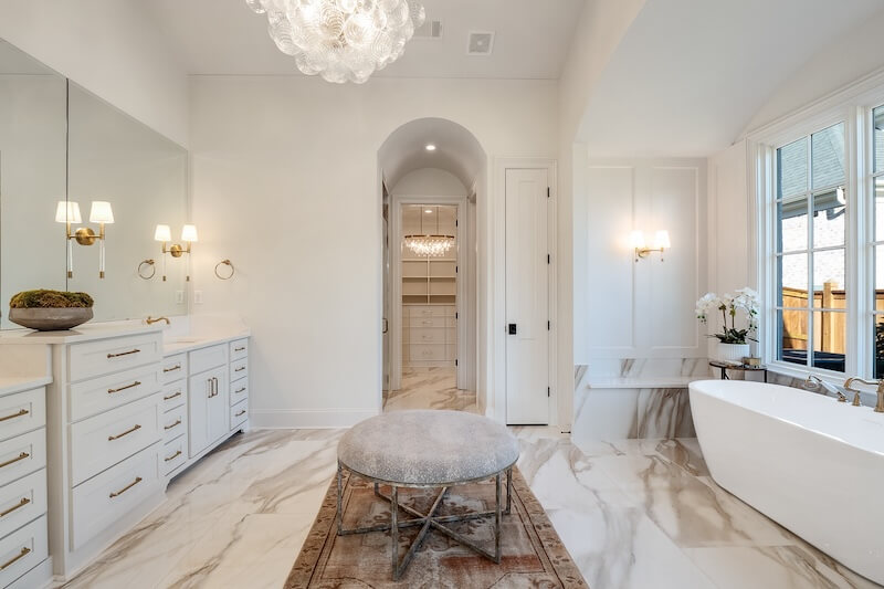 A spacious bathroom with white cabinets, a large mirror, a freestanding bathtub, a chandelier, and marble flooring. An archway leads to a walk-in closet in the background.