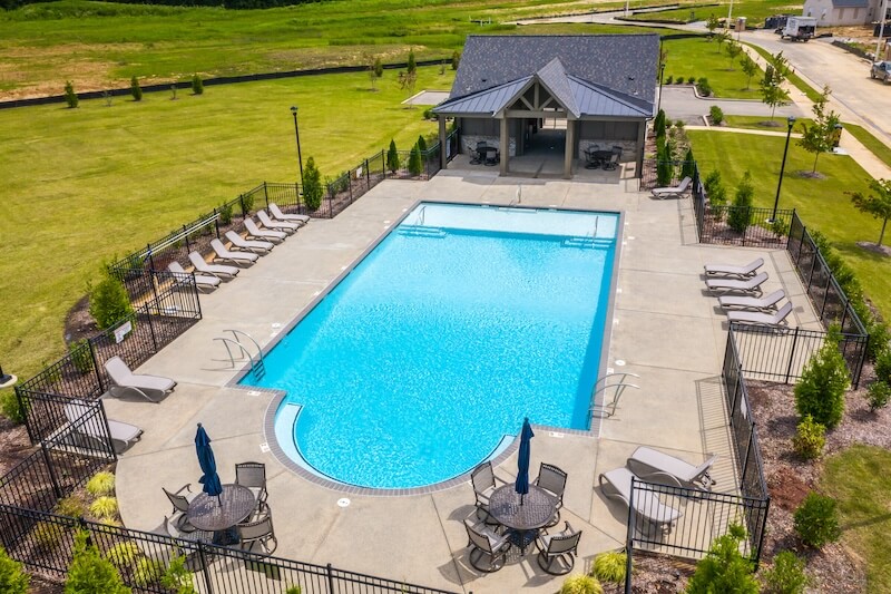 Aerial view of a rectangular outdoor pool surrounded by lounge chairs and tables with umbrellas, enclosed by a black fence. A small building with a sloped roof is located at one end of the pool area.