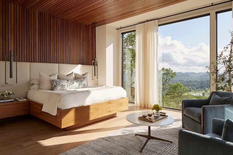A modern bedroom featuring a wooden platform bed with white bedding, large windows offering a view of rolling hills, a gray armchair, and a wooden ceiling designed by the best interior designers in Nashville.