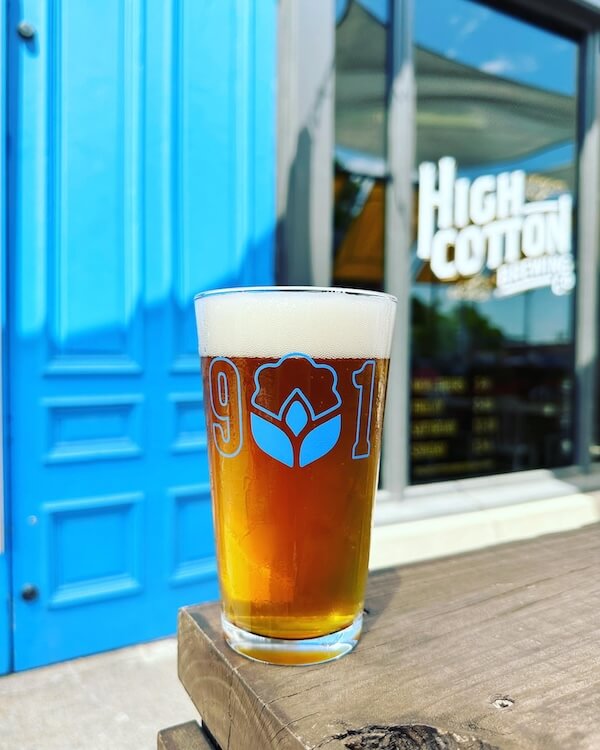 A glass of beer, featuring a "901" logo from one of the local Memphis breweries, is set on an outdoor ledge against a blue door and window with "high cotton brewing" signage.