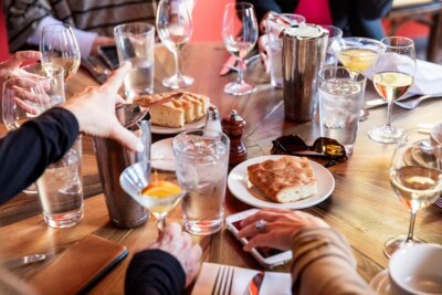 People sit around a wooden table set with plates of bread, glasses of wine and water, and various utensils, engaging in conversation and dining in the heart of Highland Park Birmingham.