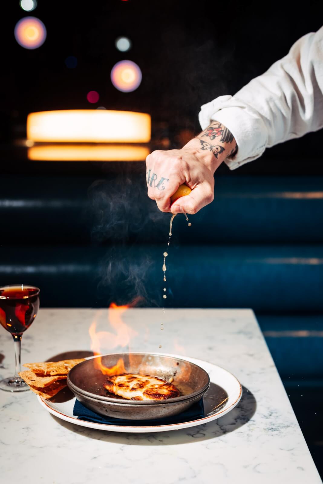 Upscale food and live music? We can get on board with that! Check out this flaming Greektown Classic Saganaki! Image: Nathan Zucker Chef squeezing lemon over a Greek saganaki that's being flambeed.