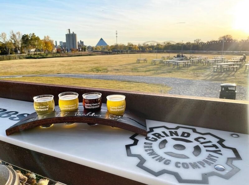 A flight of beers from local breweries on a branded tray overlooking an outdoor seating area at sunset with a cityscape and pyramid in the background.