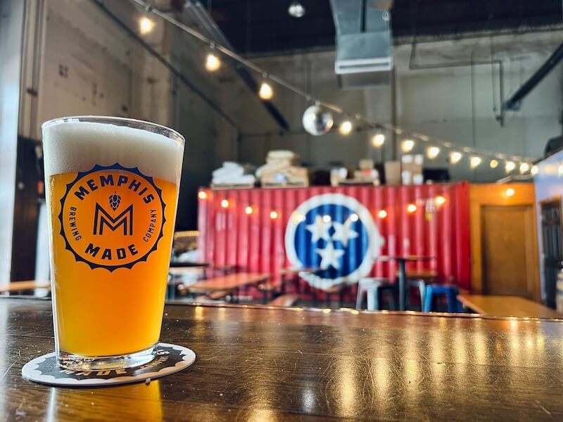 A glass of beer on a coaster with the Memphis Breweries logo, set on a bar with a vibrant red container and brewery interior in the background.
