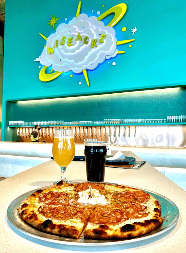 A fresh pepperoni pizza with a dollop of cream on a metal tray, flanked by a glass of pale beer from Memphis Breweries and a dark beverage, with a colorful "wiseacre