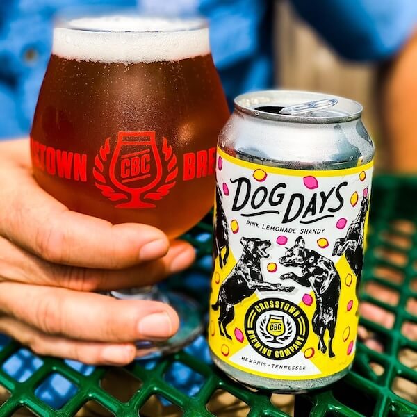 A person holding a glass of beer next to a colorful beer can with the label "dog days pink lemonade shandy" from Crosstown Brewing Company.
