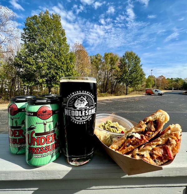 Craft beer from local Memphis breweries and a loaded hot dog on an outdoor table against a backdrop of trees and a parking lot.