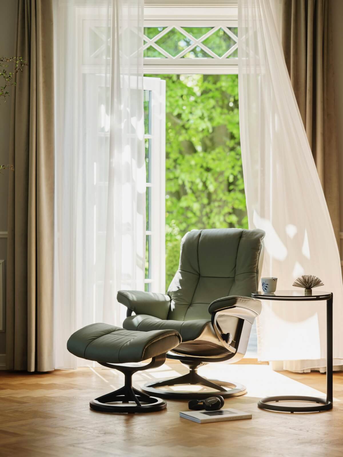 A cozy armchair with a matching footrest sits beside a round side table with a mug, book, and headphones. Sunlight filters through the open window and sheer curtains, revealing greenery outside—a perfect example of Scandinavian design's harmony showcased in this serene nook.