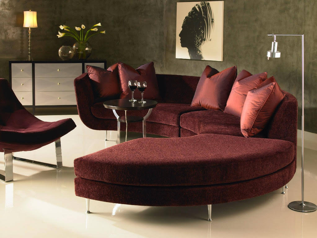 Modern living room with a curved maroon sofa, matching chair, and pillows. A round side table with wine glasses complements the black and white cabinet. The floral artwork and floor lamp add a touch of Scandinavian Design elegance, creating a space worthy of any Design Gallery.