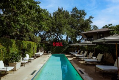 A narrow outdoor pool is lined with lounge chairs and umbrellas, surrounded by greenery. Showcasing unique hotel amenities, the space features the word “SOUL” in red letters on the far end wall, creating a vibrant and inviting atmosphere.