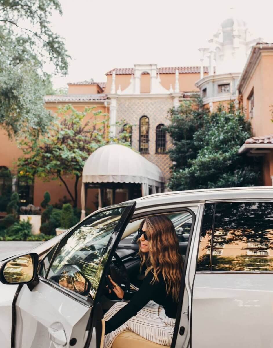 Woman wearing sunglasses steps out of a white car in front of an ornate building with arched windows and a domed entrance, surrounded by trees and greenery, ready to experience the luxurious hotel amenities awaiting inside.