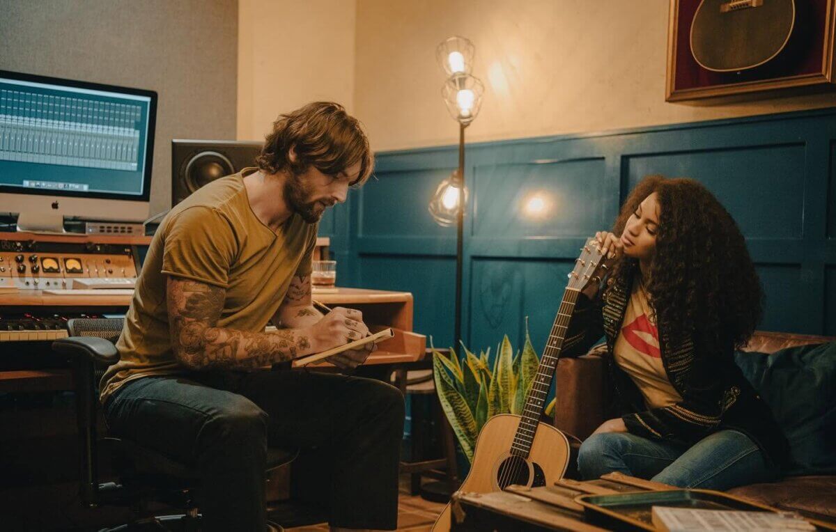 A man writes on a notepad at a studio desk while a woman with a guitar sits nearby, listening thoughtfully. Studio equipment and a computer monitor are visible in the background, creating an atmosphere as inspiring as unique hotel amenities.