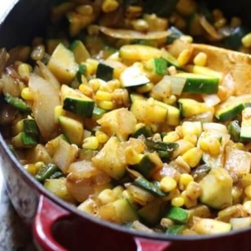 A red pot brimming with a Mexican vegetable side dish, where zucchini, corn, onions, and spices meld harmoniously. A wooden spoon awaits to serve this delightful medley.