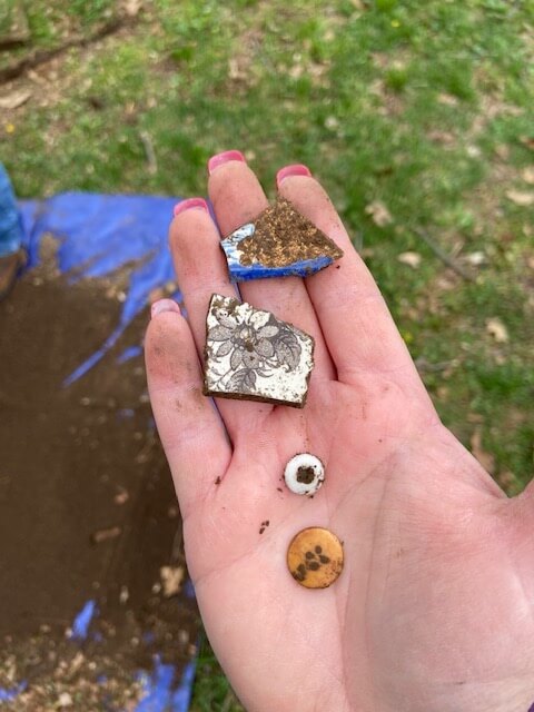Rachael's hand holding buttons and dishware pieces she pulled from the ground.