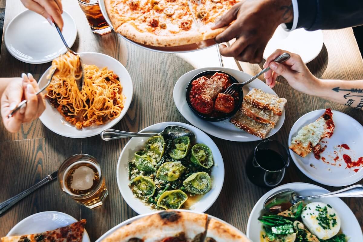 A table at The Nations Nashville showcases a variety of Italian dishes, including pizza, spaghetti, stuffed pasta shells, and meatballs with bread. Hands reach eagerly for the delicious offerings and refreshing beverages.