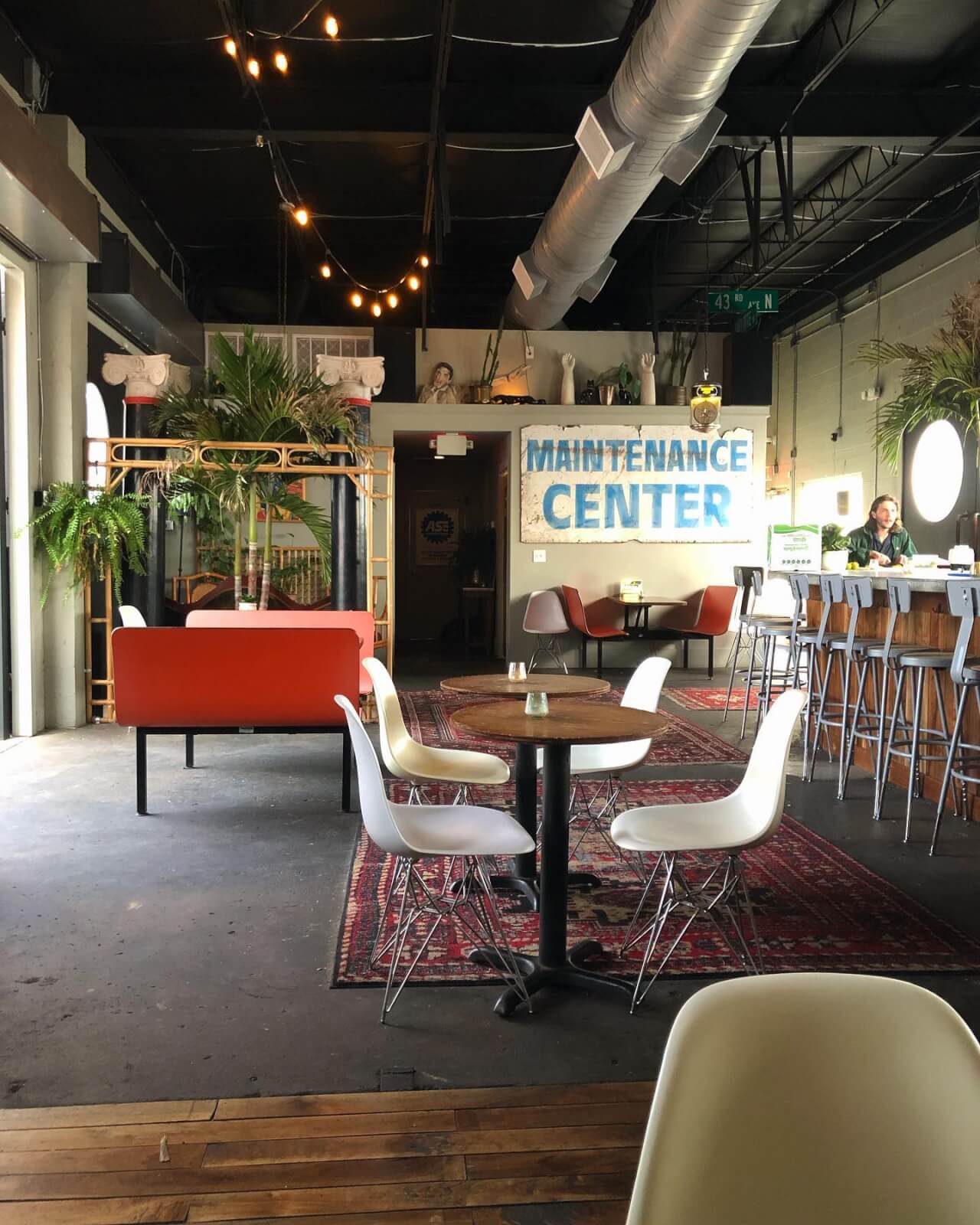 Café interior labeled "Maintenance Center" in the trendy Nations Nashville area, featuring white chairs, wooden tables, a red couch, and lush plants. A bar with stools stands to the right under inviting overhead lights as someone works on a laptop.