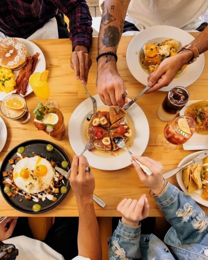 The Best Brunches in Louisville: 13 Must-Try Spots - 5 Four people enjoying the best brunch in Louisville, sharing pancakes with fruit. Various dishes like eggs, toast, bacon, and drinks are arranged on a wooden table for a cozy meal together.