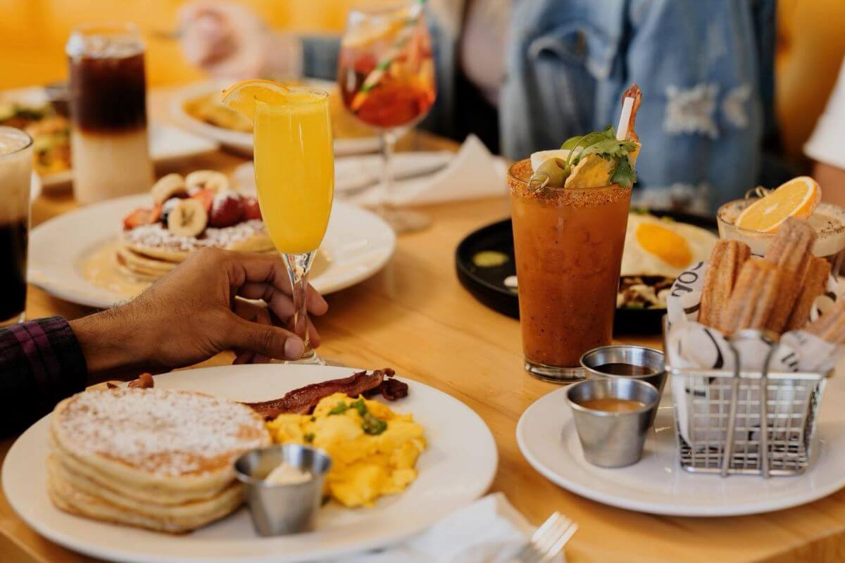 A table set with plates of pancakes, scrambled eggs, bacon, churros, and drinks like orange juice and a cocktail, with people gathered around—classic signs of enjoying the best brunch in Louisville.