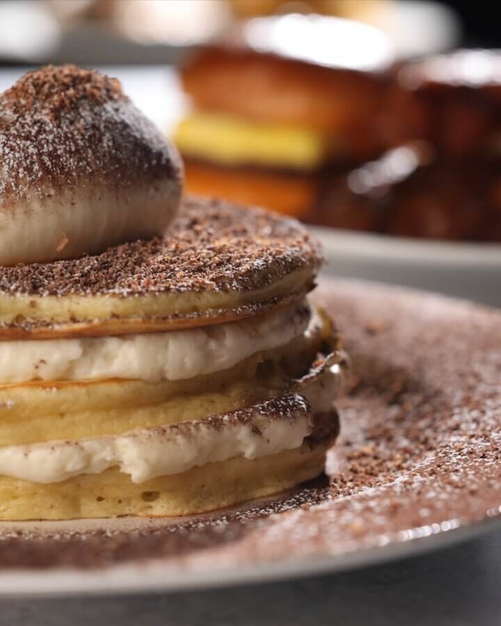The Best Brunches in Louisville: 13 Must-Try Spots - 2 A stack of pancakes filled with cream and topped with whipped cream and cocoa powder, showcased as part of the best brunch in Louisville, with other breakfast foods blurred in the background.