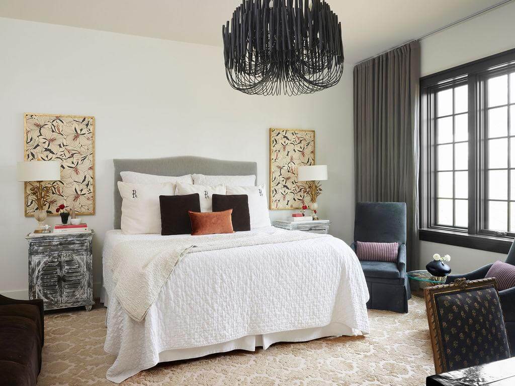 The Top Interior Designers and Architects in Birmingham - 9 Bedroom crafted by some of the best interior designers and architects in Birmingham, featuring a large bed with white bedding and decorative pillows. Two bedside tables with lamps, floral artwork, a dark chandelier, and a large window draped with gray curtains complete the decor.