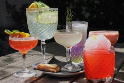 A variety of colorful cocktails and a small pastry on a wooden table outdoors in Forest Park, with sunlight highlighting the drinks.