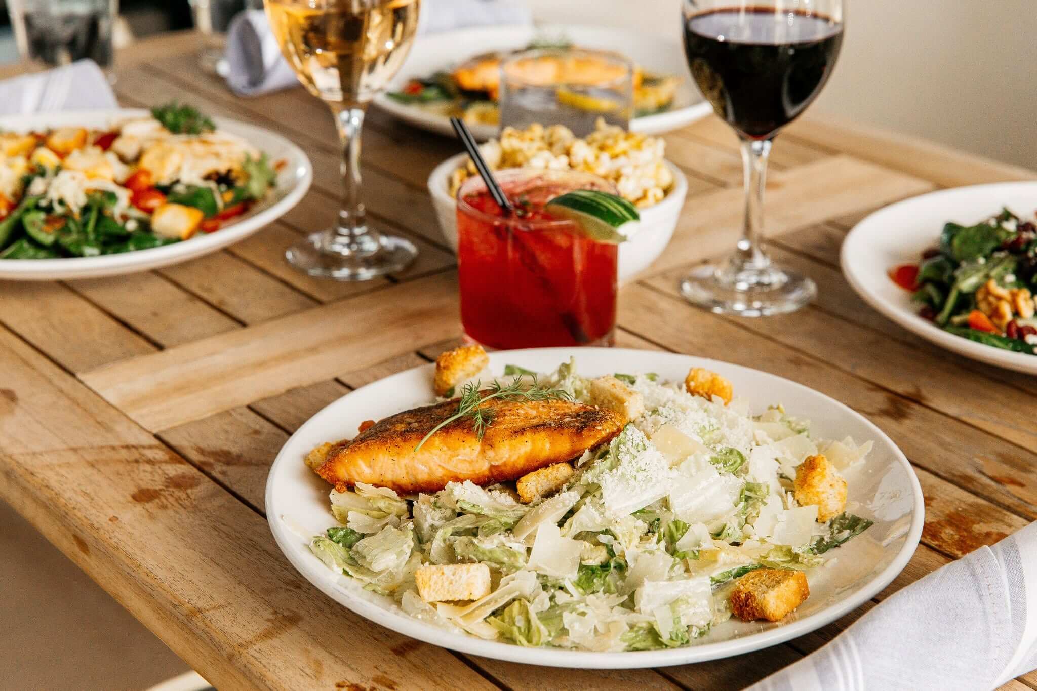 Plate of Caesar salad with salmon, surrounded by salads, drinks, and bowls of food on a wooden table in the heart of South Main Memphis.