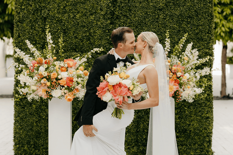 The Top Memphis Florists + Floral Designers - 3 Bride and groom kissing in front of greenery wall and large floral arrangements