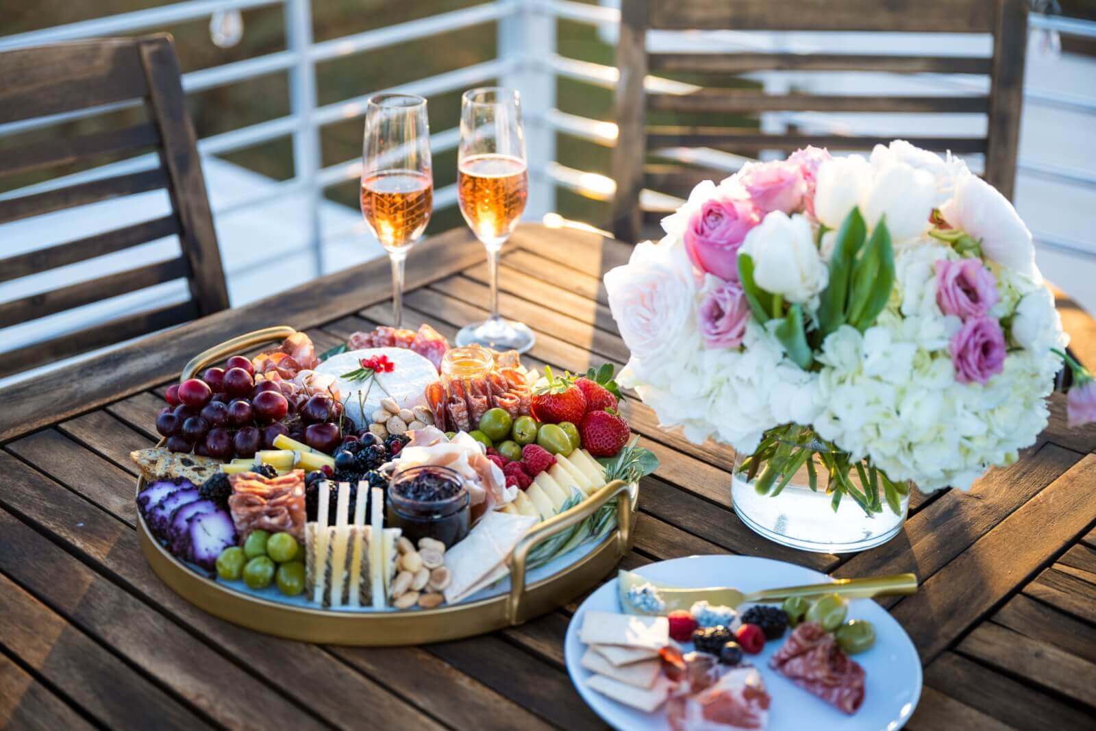 A wooden table is artfully arranged by the best event planners, showcasing a charcuterie board with cheeses, fruits, and nuts alongside two glasses of rosé and a white and pink floral arrangement.