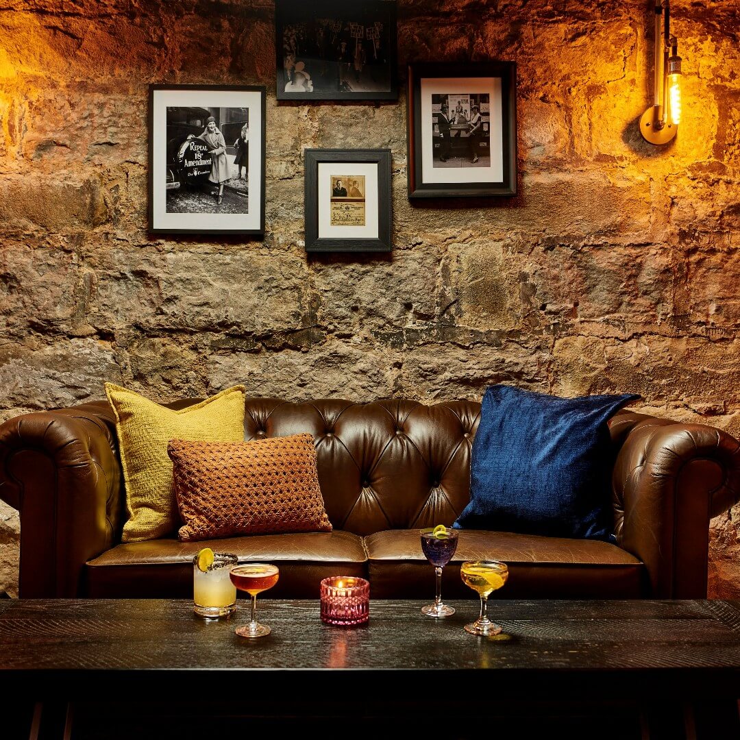 Brown leather couch with three pillows, next to a table with cocktails.