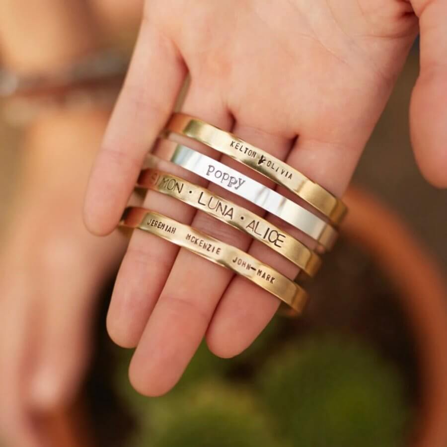 A hand holding several engraved golden bracelets with names like "Kelton," "Olivia," "Poppy," "Luna," and "Alice"