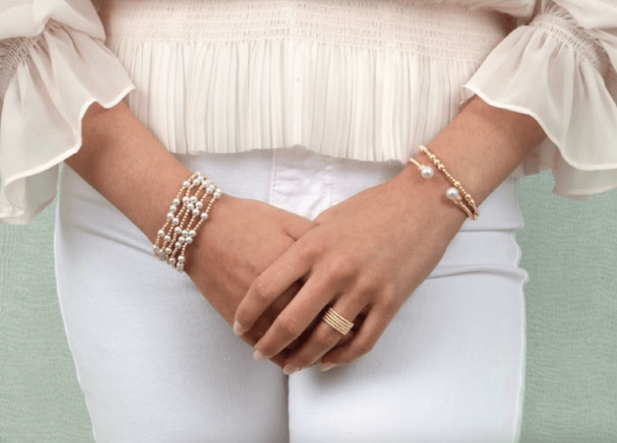 Close-up of a woman's hands clasped over her lap, adorned with several bracelets, wearing a white blouse and white pants.
