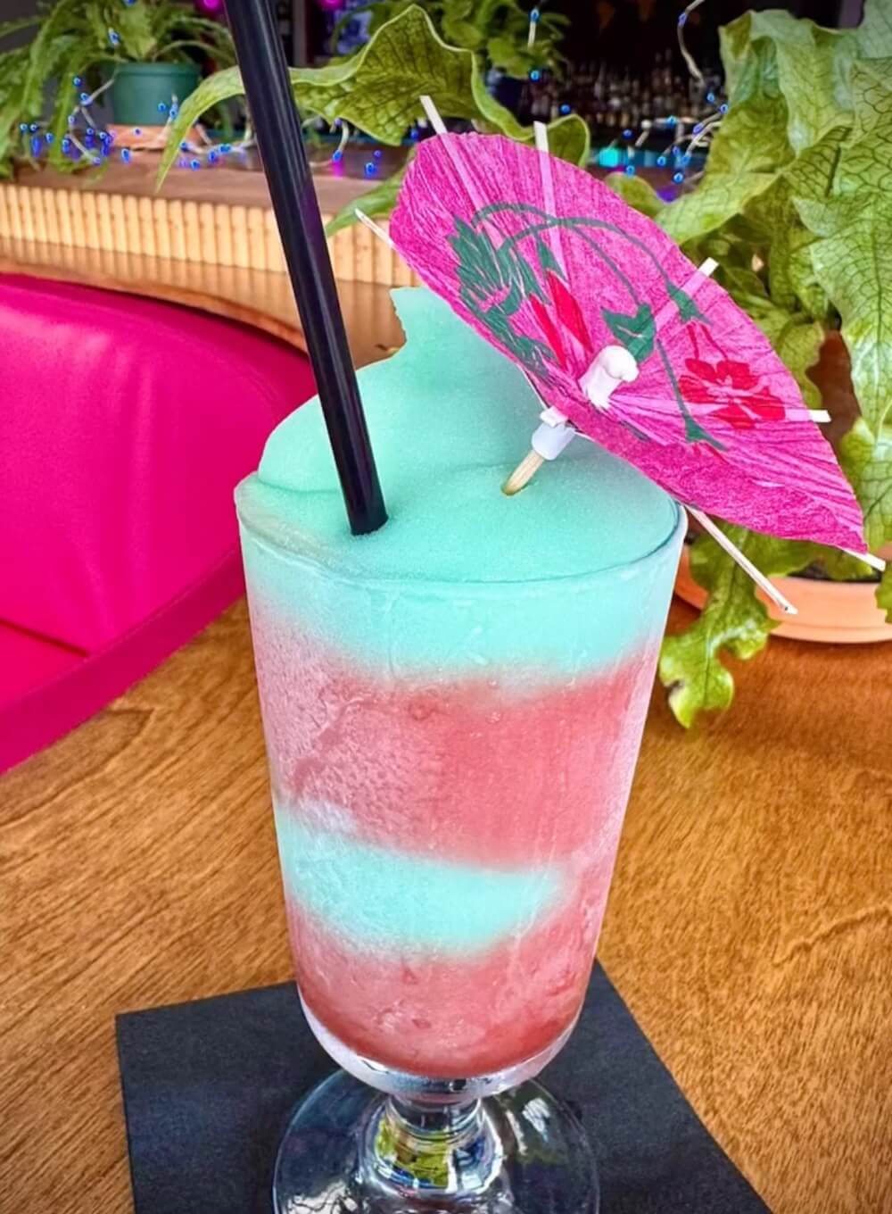 A layered frozen cocktail with blue and pink slush, served in a tall glass with a straw and a decorative pink paper umbrella, placed on a black napkin on a wooden table.