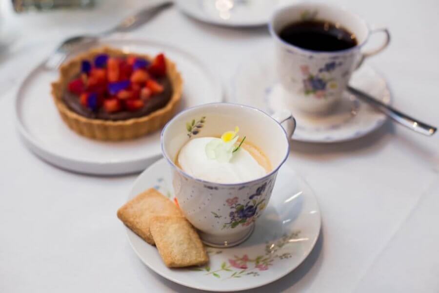 Cup of cappuccino with biscuits, a chocolate tart with berries on a plate, and a cup of black coffee set on a white tablecloth evoke the essence of an elegant afternoon tea in Birmingham.
