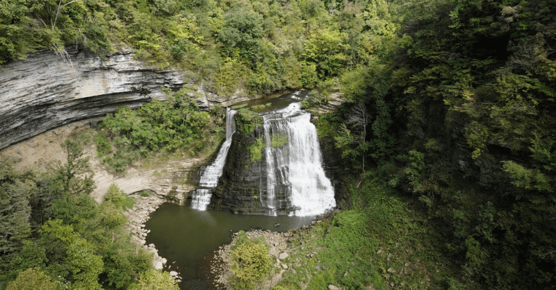 Aerial view of one of the Best Waterfalls in Tennessee, cascading over layered rock cliffs into a small pool, surrounded by dense, green forest.