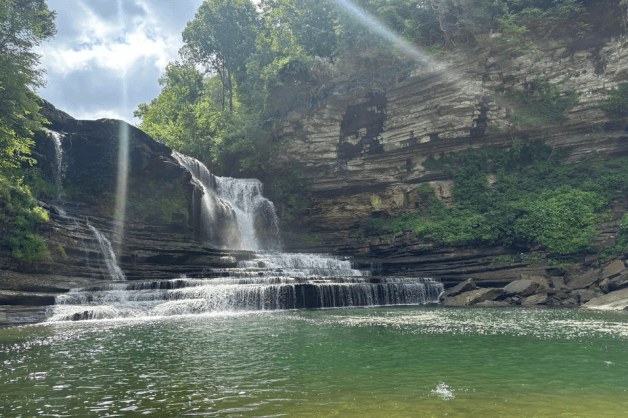 Waterfall cascading over layered rock formations into a green pool, surrounded by lush greenery and sunlight streaming through trees—one of the best waterfalls in Tennessee for nature lovers to explore.
