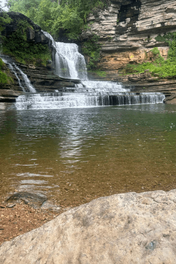 A multi-tiered waterfall, considered one of the Best Waterfalls in Tennessee, flows into a clear, shallow pool surrounded by rocky ledges and lush green vegetation.