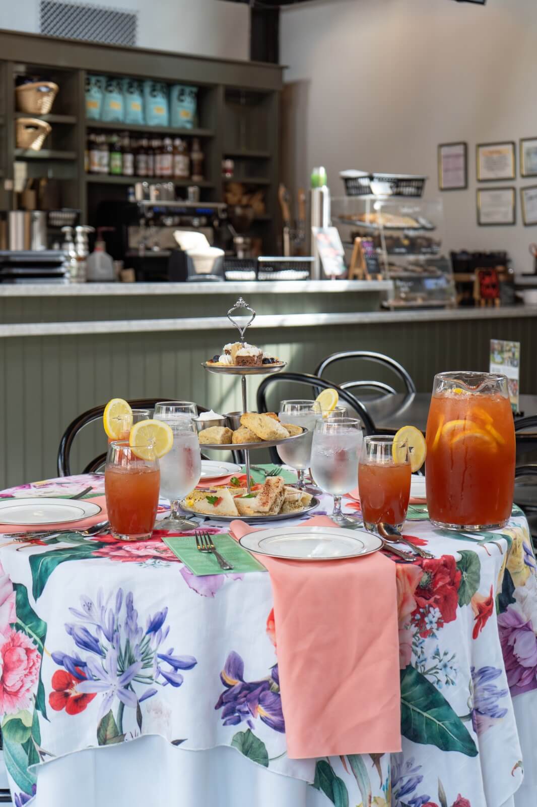 A floral tablecloth covers a round table set with tea service—iced tea, water, a pitcher of tea, scones, and pastries—in a café with a counter in the background.