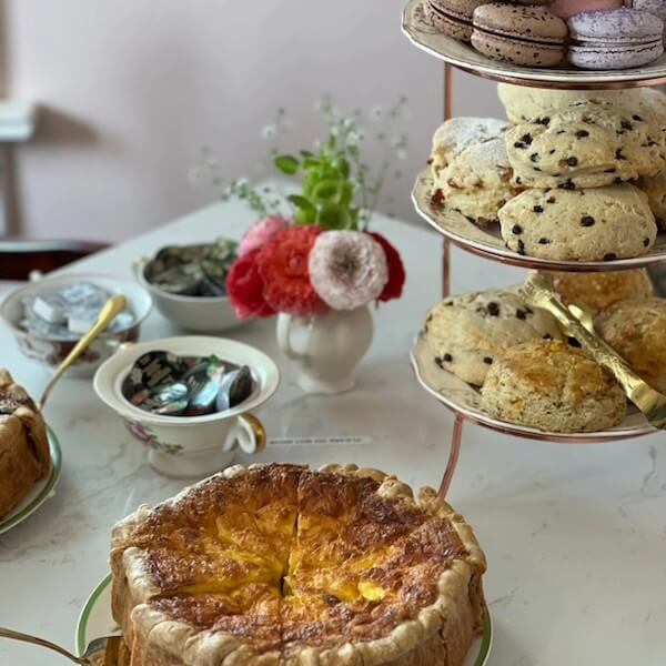 A table set for tea service features a savory pie, a tiered stand with chocolate chip scones and macarons, bowls of jam, and a small vase of flowers.