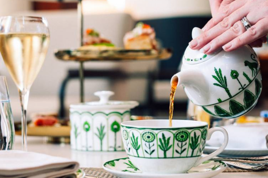 A hand pours tea from a white teapot with green floral designs into a matching teacup, capturing an elegant tea service; pastries and a glass of champagne are in the background.