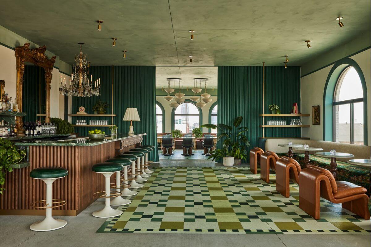 A stylish NuLu bar and lounge with green and brown decor, marble tables, leather chairs, a patterned rug, and large arched windows letting in natural light.